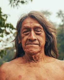 Ancient Indigenous man with long hair and weathered skin, outdoors in natural setting. 