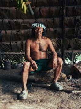 Maori elder sitting outdoors, wearing traditional feathered headgear, in a cultural setting. 