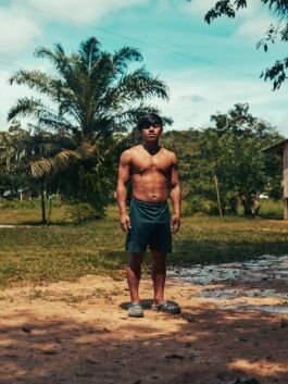Young boy standing shirtless outdoors in a lush, tropical environment with palm trees and greenery. 