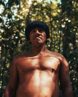 Indigenous man standing shirtless in a forest with sunlight filtering through trees. 