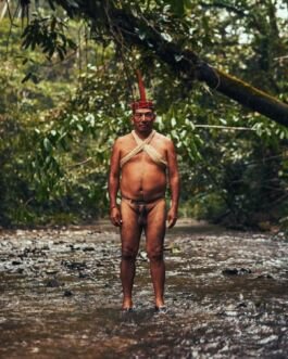 Indigenous man in traditional attire standing in a river surrounded by lush forest, showcasing cultu. 