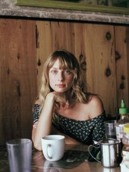 Contemplative woman with blonde hair and floral off-shoulder top sitting at cafe table, surrounded b. 