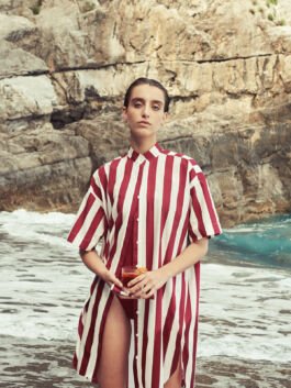 Person wearing a red-and-white striped shirt dress stands at the rocky seaside, holding a glass drink. 