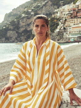 Woman seated on a pebble beach wearing a yellow-and-white striped dress, with a cliffside town in the background. 
