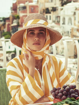 Woman wearing a wide-brim straw hat and yellow‑striped blouse sits at an outdoor cafe, with grapes on the table. 