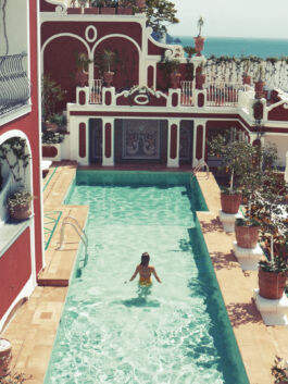Woman swimming in a turquoise rectangular pool surrounded by ornate red-and-white Mediterranean architecture and potted plants; sea in the background. 