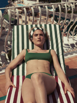 Woman in a green bikini sunbathing on a striped lounge chair on a balcony overlooking a coastal town. 