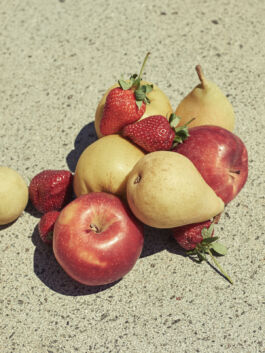 Pile of fresh fruit on a rough concrete surface: red apples, yellow pears, and strawberries intermingled. 
