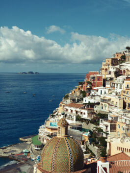 Colorful hillside town along the coast with a tiled mosaic dome in the foreground and blue sea beyond. 