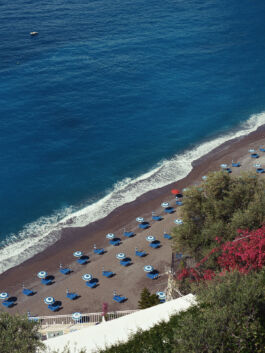 Pebble beach lined with blue sun loungers and white-blue umbrellas along the shoreline, blue sea beyond, with a small boat in the distance and pink flowering bushes in the foreground. 