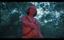 Shirtless man wearing a headband holds a long spear wrapped with rope in a forest at dusk. 