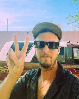 Robin Mellor enjoying a sunny day, making a peace sign, wearing sunglasses and a cap outdoors. 